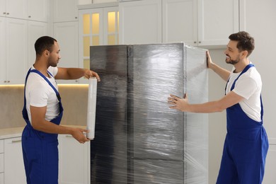 Photo of Male movers with stretch film wrapping refrigerator in new house