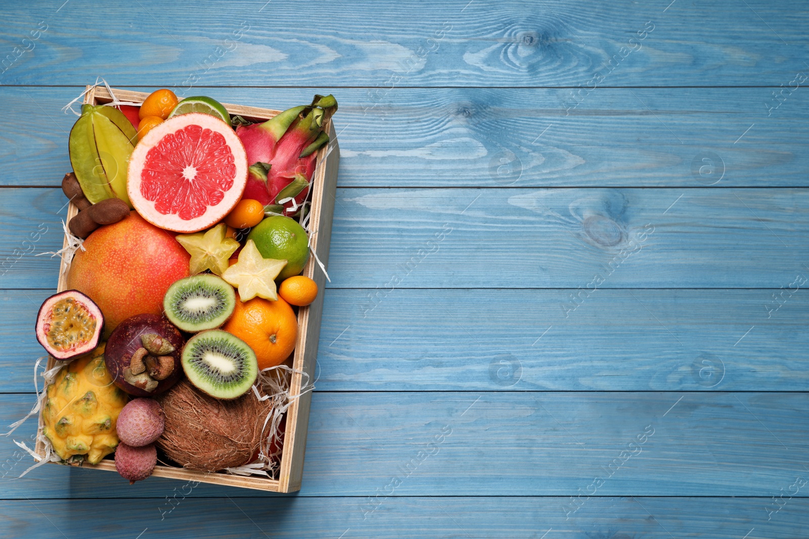 Photo of Different tropical fruits in box on blue wooden background, top view. Space for text