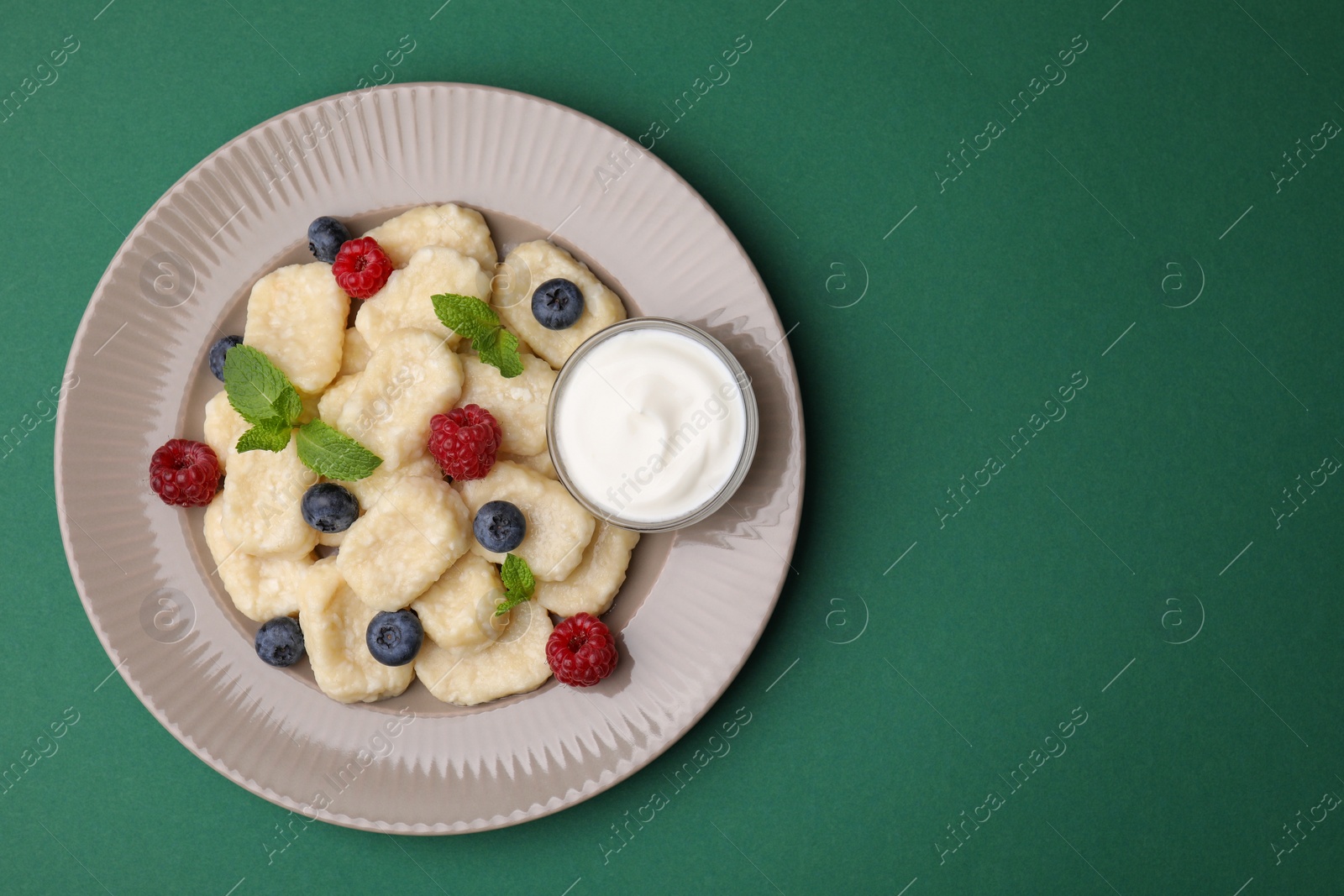 Photo of Plate of tasty lazy dumplings with berries, mint leaves and sour cream on dark green background, top view. Space for text