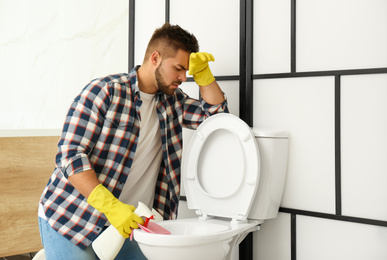 Photo of Young man feeling disgust while cleaning toilet bowl in bathroom