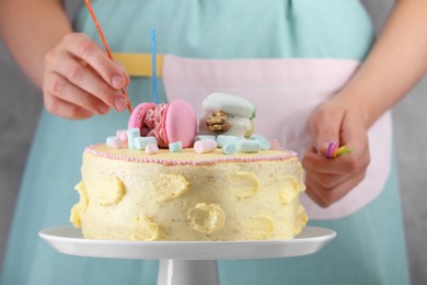 Photo of Woman putting candles on cake decorated with macarons and marshmallows on blurred background, closeup