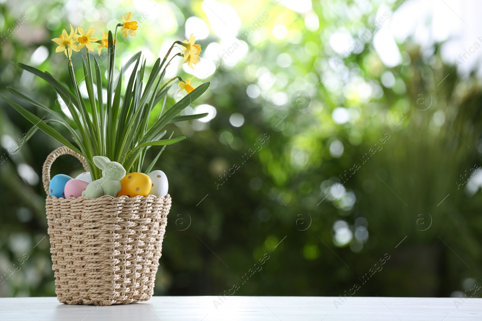 Photo of Easter bunny toy, dyed eggs and flowers in wicker basket on white wooden table against blurred green background. Space for text