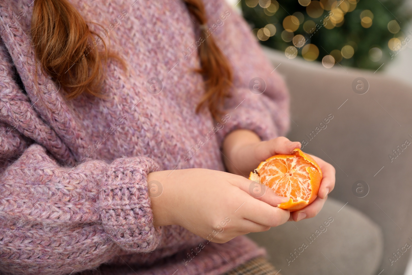 Photo of Little girl peeling fresh tangerine on sofa at home, closeup