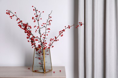 Photo of Hawthorn branches with red berries in vase on wooden table indoors, space for text