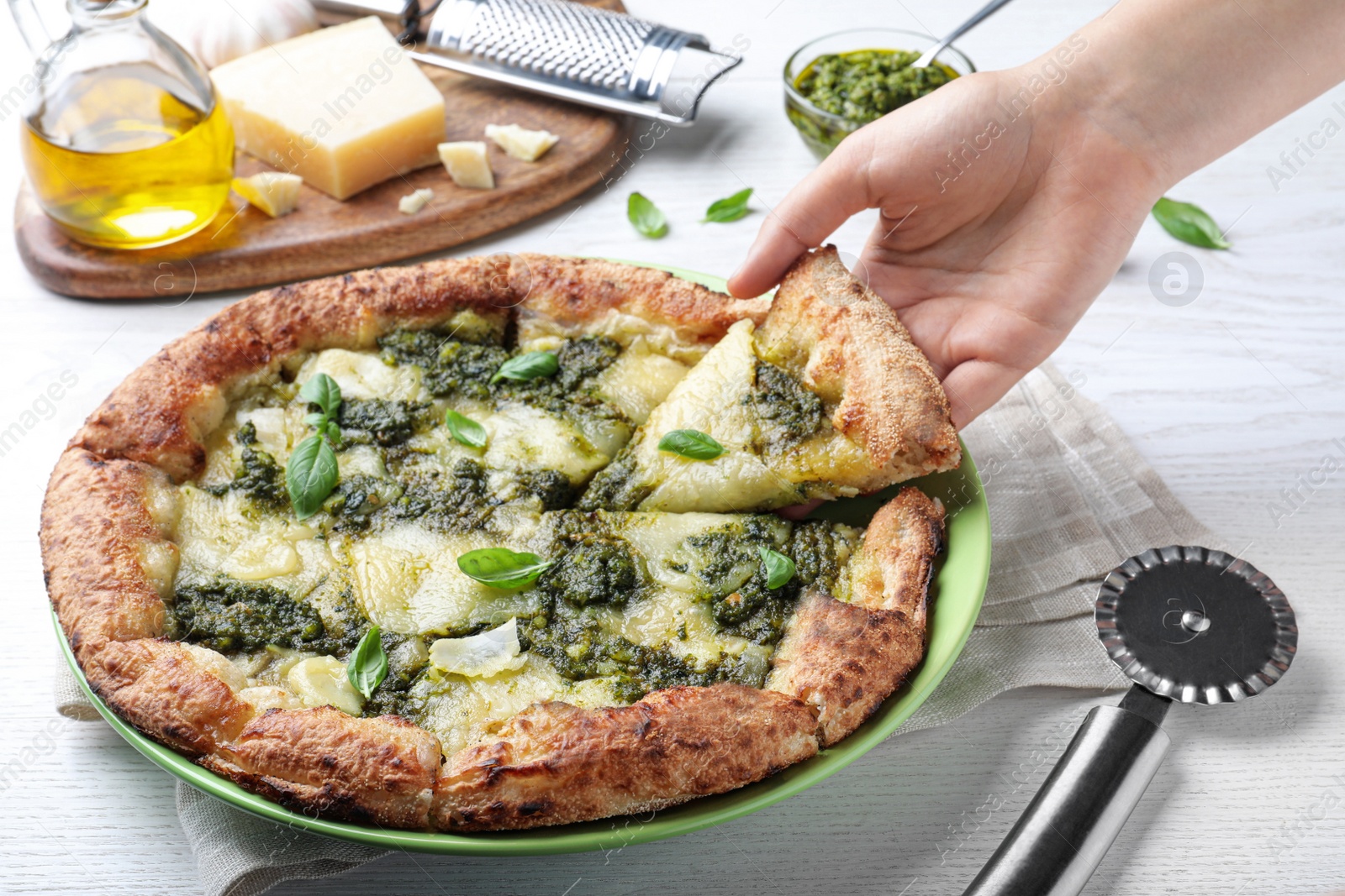 Photo of Woman taking slice of delicious pizza with pesto, cheese and basil at white wooden table, closeup
