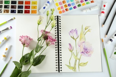 Photo of Painting of eustomas in sketchbook, flowers and art supplies on white background, top view