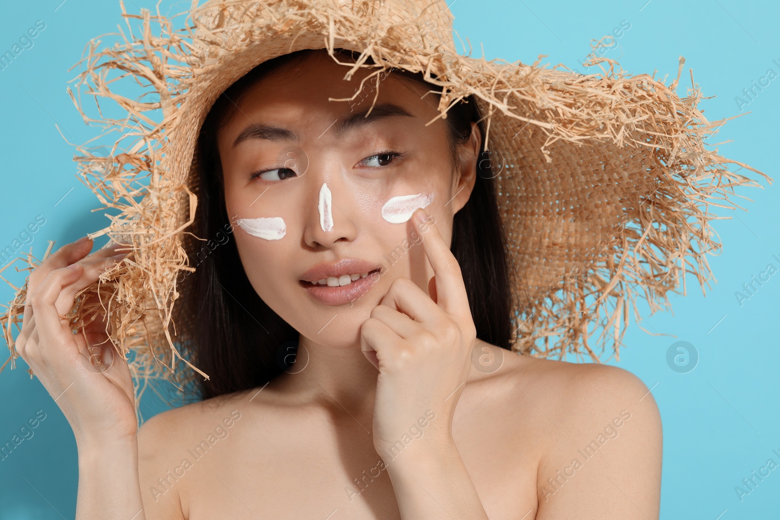 Photo of Beautiful young woman in straw hat with sun protection cream on her face against light blue background