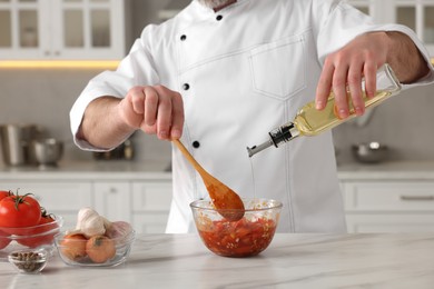 Photo of Professional chef pouring oil from bottle into delicious tomato sauce at white marble table indoors, closeup