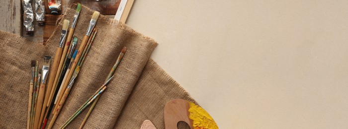 Photo of Natural burlap fabric and different painting supplies on beige background, top view