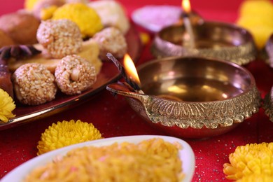 Diwali celebration. Diya lamps and tasty Indian sweets on shiny red table, closeup