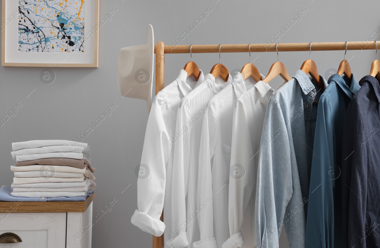 Photo of Rack with different stylish shirts, hat and chest of drawers near grey wall in room. Organizing clothes
