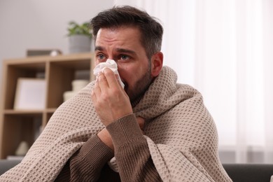 Sick man wrapped in blanket with tissue blowing nose on sofa at home. Cold symptoms