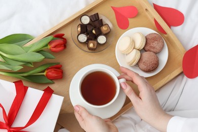 Photo of Tasty breakfast served in bed. Woman with tea, desserts, gift box and flowers at home, top view