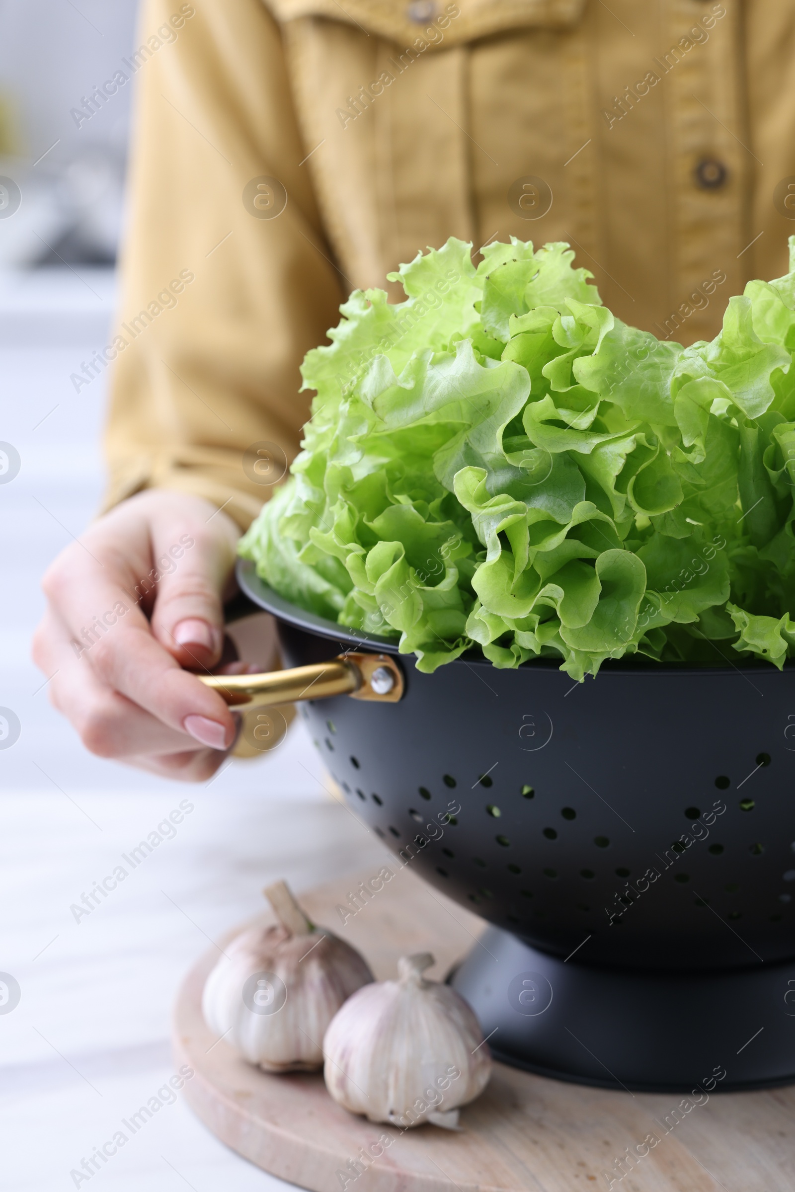 Photo of Woman holding black colander with lettuce at white table indoors, closeup