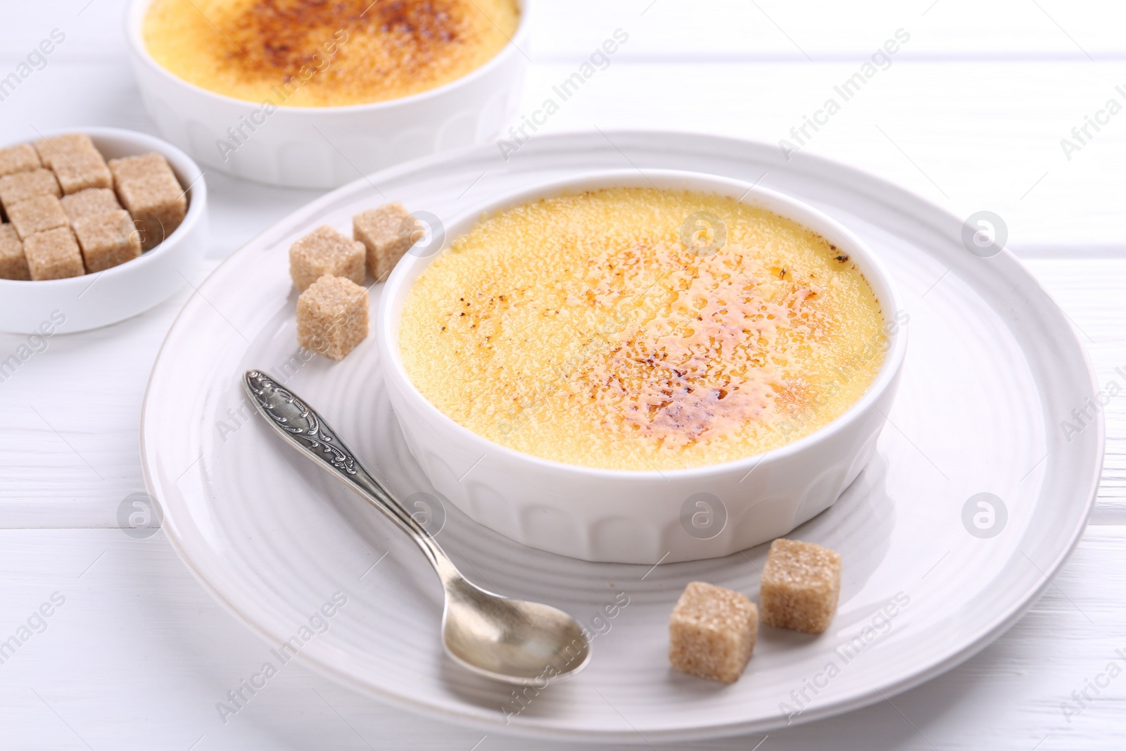 Photo of Delicious creme brulee in bowls, sugar cubes and spoon on white wooden table
