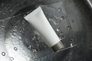 Photo of Moisturizing cream in tube on glass with water drops against metal background, top view