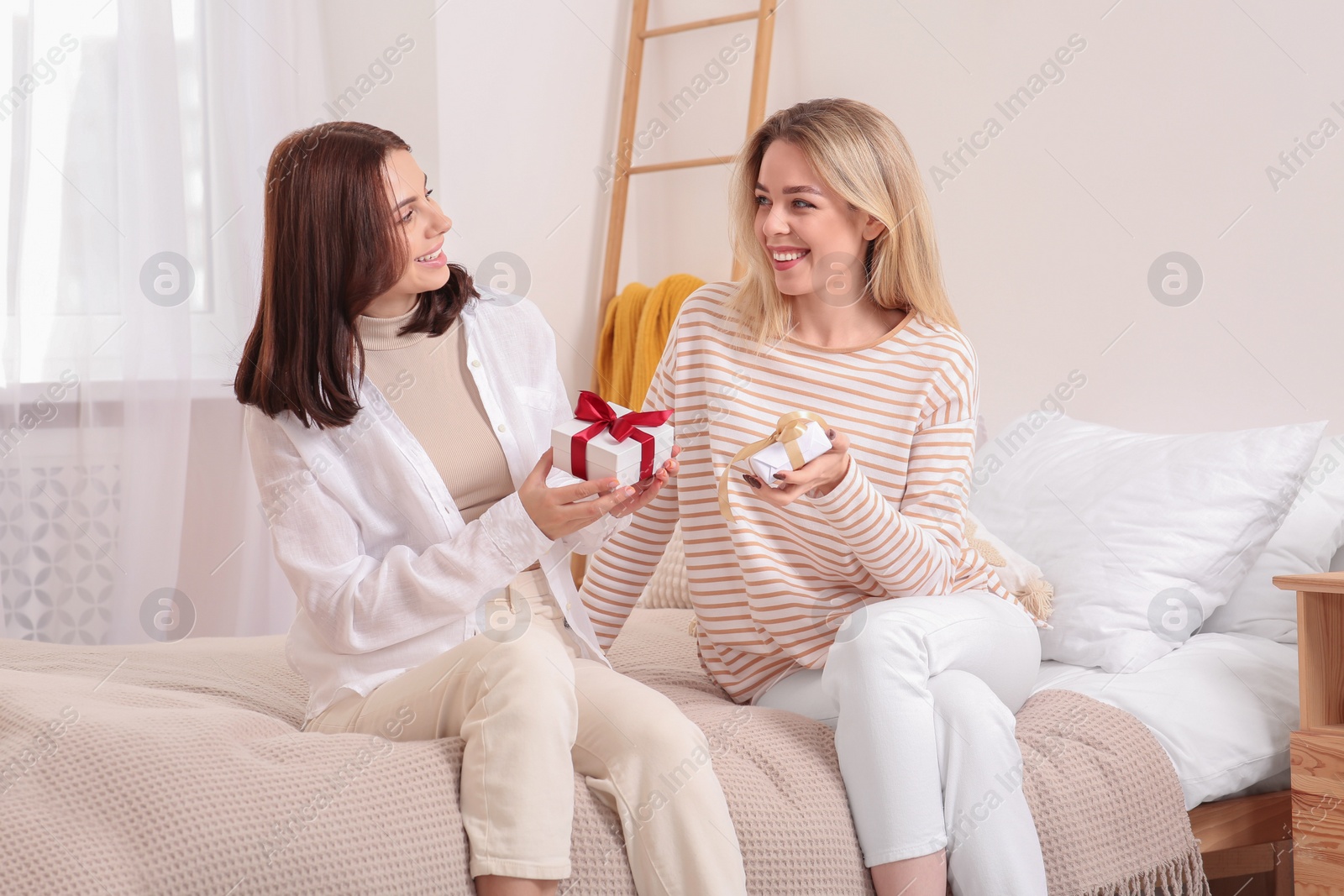 Photo of Smiling young women presenting gifts to each other on bed at home