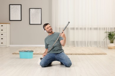 Photo of Enjoying cleaning. Happy man in headphones with mop singing while tidying up at home