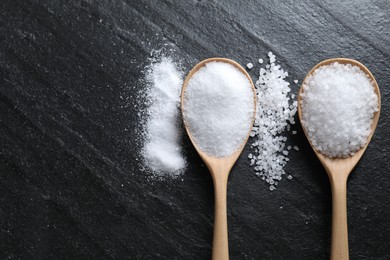 Photo of Organic salt in spoons on black table, flat lay. Space for text