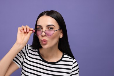 Photo of Beautiful young woman in stylish sunglasses blowing kiss on purple background. Space for text