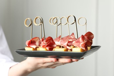 Woman holding plate of tasty canapes with ham and cheese indoors, closeup