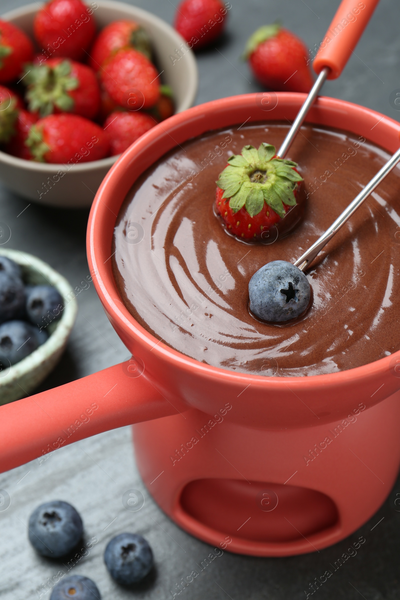 Photo of Dipping fresh berries in fondue pot with melted chocolate at grey table, above view