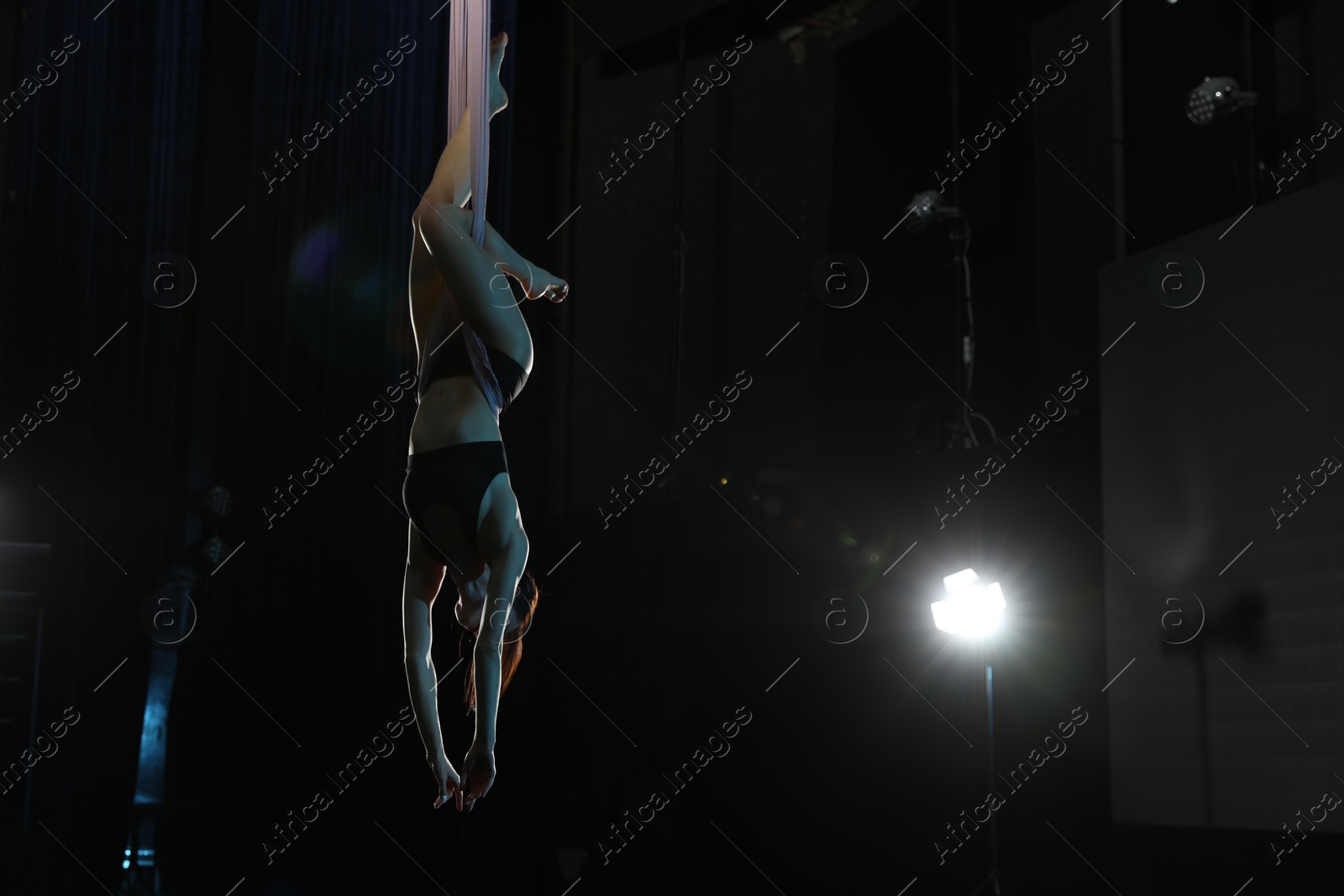 Photo of Young woman performing acrobatic element on aerial silk against dark background. Space for text