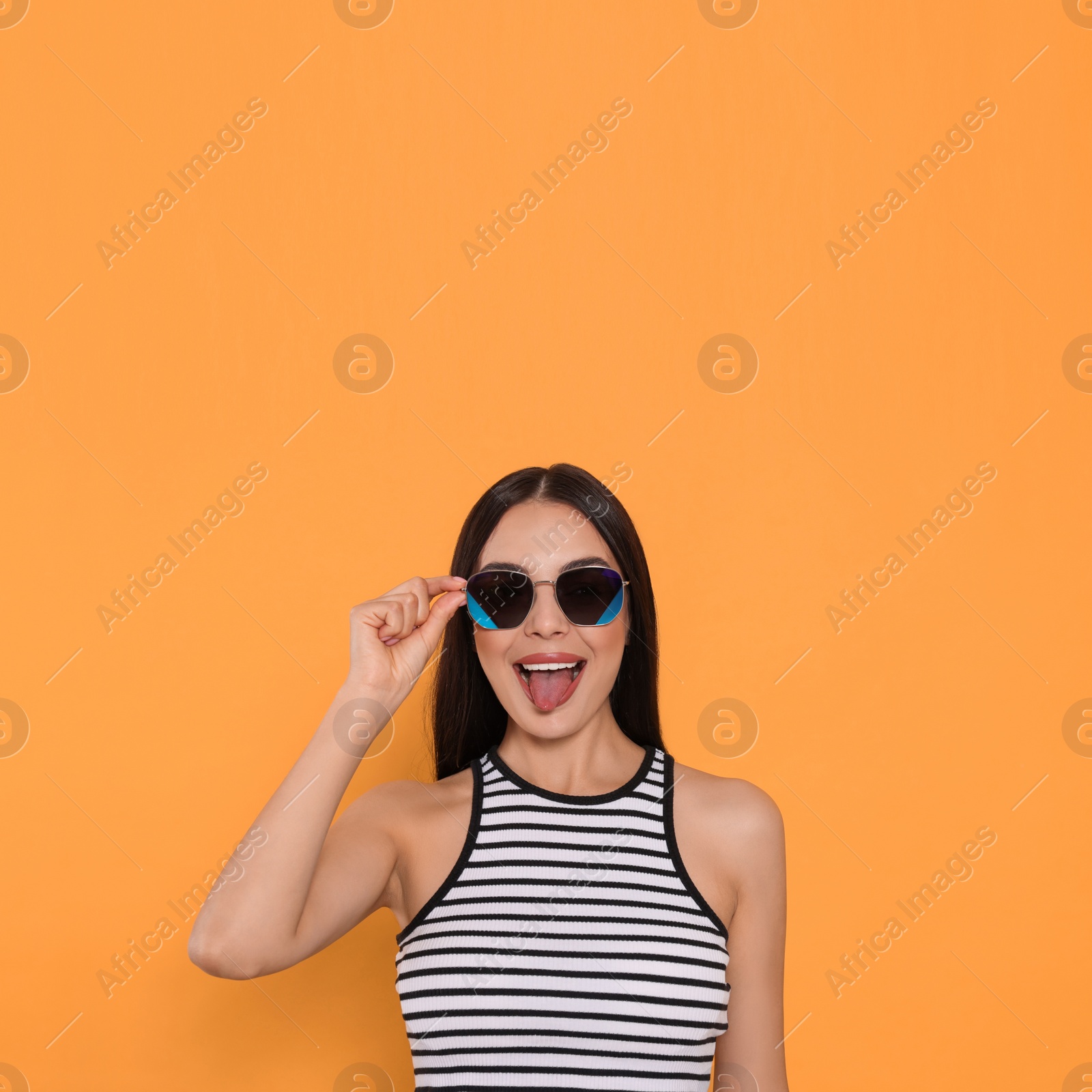 Photo of Attractive happy woman in fashionable sunglasses showing tongue against orange background. Space for text