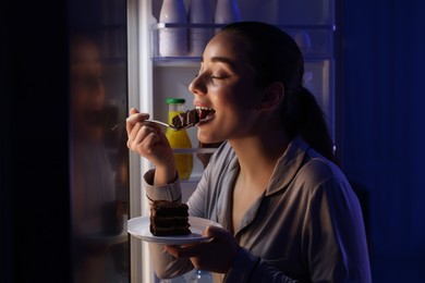 Photo of Young woman eating cake near refrigerator in kitchen at night. Bad habit