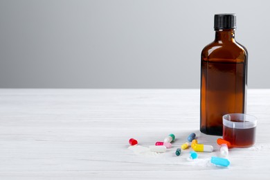 Photo of Pills, bottle with measuring cup of syrup on wooden table against white background, space for text. Cough and cold medicine