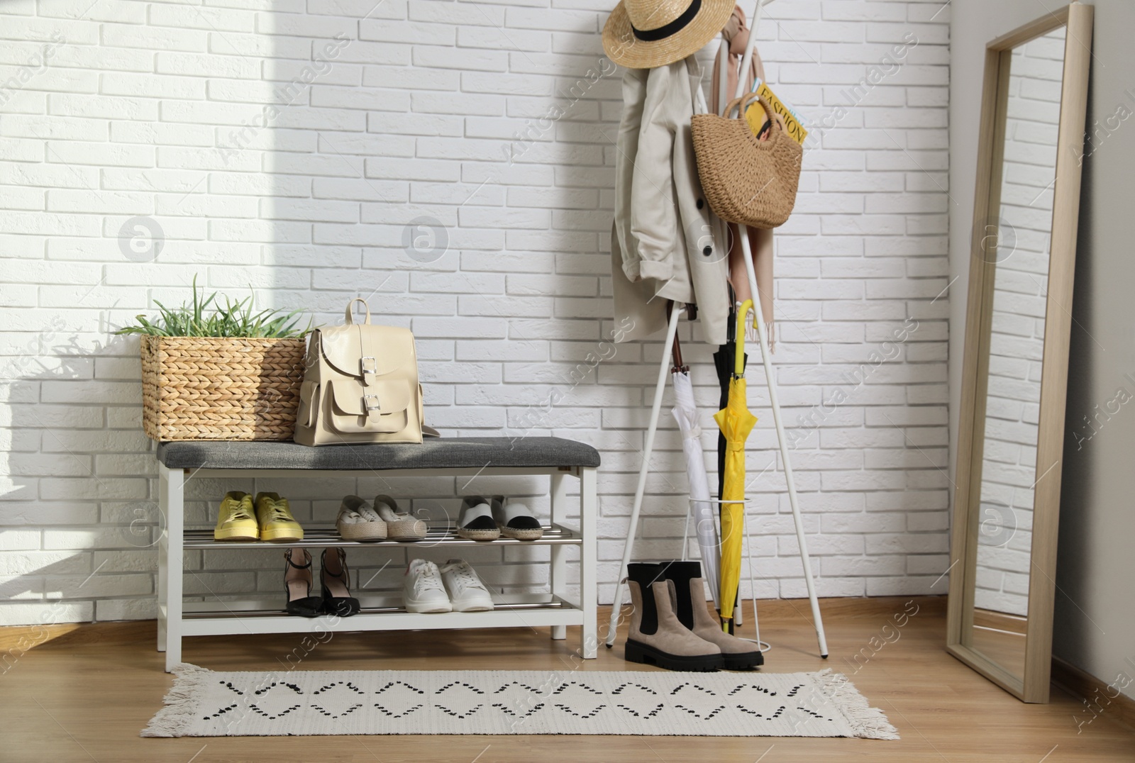 Photo of Stylish hallway interior with coat rack, shoe storage bench and mirror near white brick wall