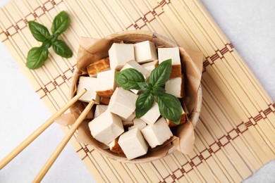 Photo of Bamboo mat with bowl of smoked tofu cubes and basil on white table, flat lay