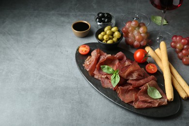 Photo of Delicious bresaola, grissini sticks, olives, tomato and grapes on grey textured table. Space for text