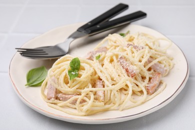 Photo of Plate of tasty pasta Carbonara with basil leaves, fork and spoon on white tiled table, closeup