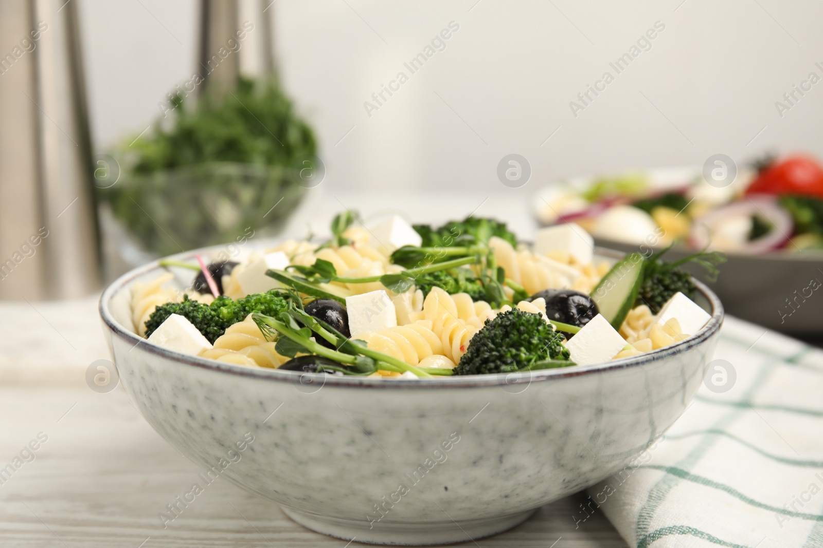 Photo of Bowl of delicious pasta with cucumber, olives, broccoli and cheese on white wooden table, closeup