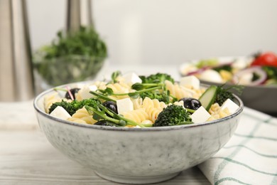 Bowl of delicious pasta with cucumber, olives, broccoli and cheese on white wooden table, closeup