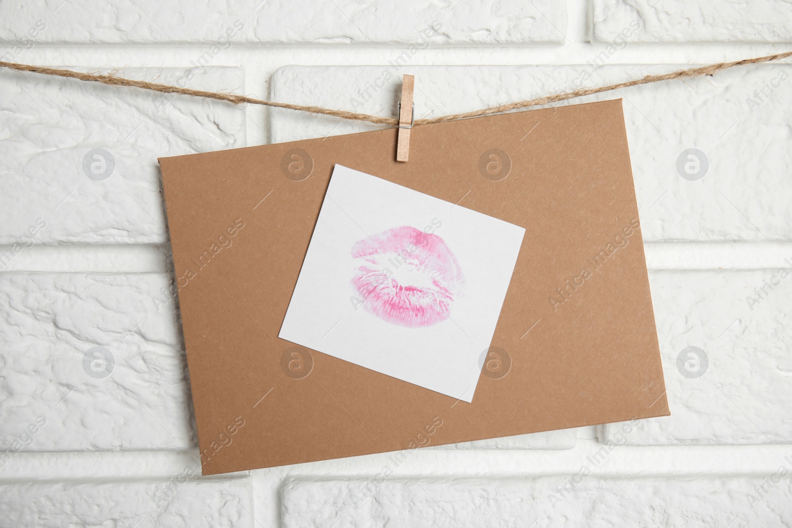 Photo of Envelope and card with lip print hanging on twine near white brick wall. Love letter