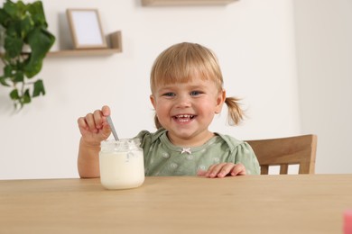 Photo of Cute little child eating tasty yogurt with spoon from jar at wooden table