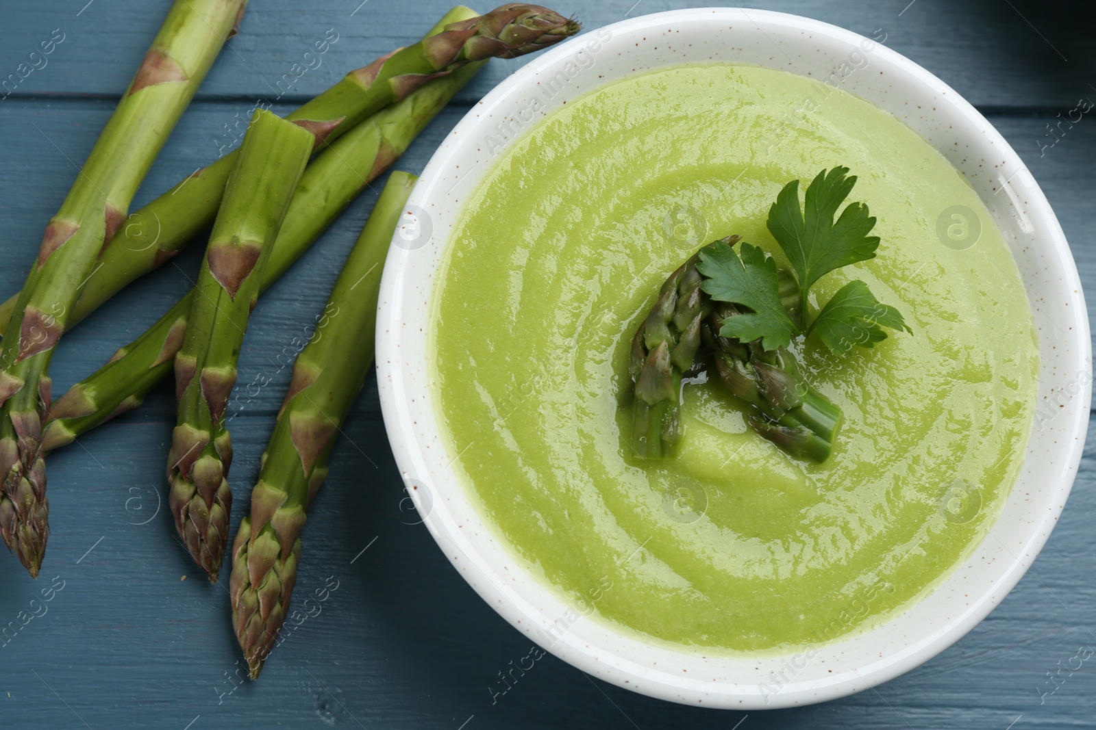 Photo of Delicious asparagus soup served on blue wooden table, flat lay