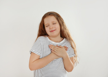 Cute grateful little girl with hands on chest against light background