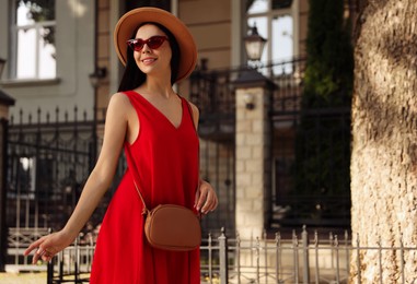 Photo of Beautiful young woman with stylish bag in red dress near building outdoors, space for text