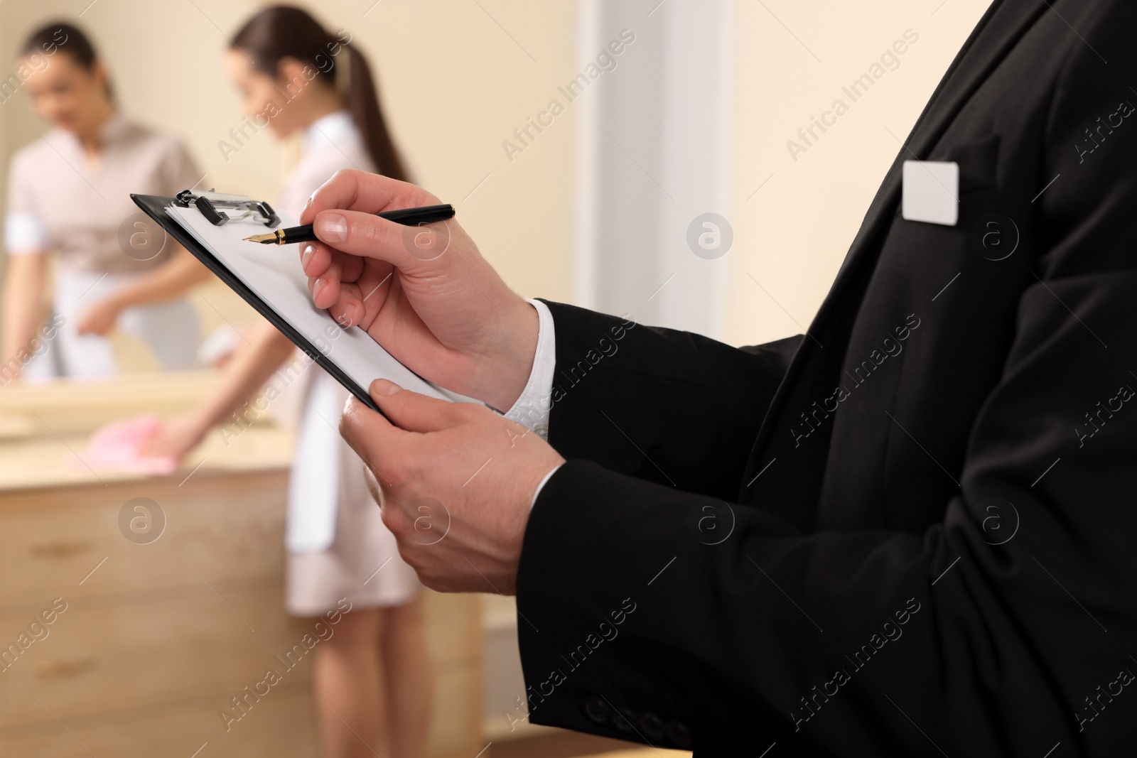 Photo of Man wearing suit with clipboard checking maid's work in hotel room, closeup. Professional butler courses