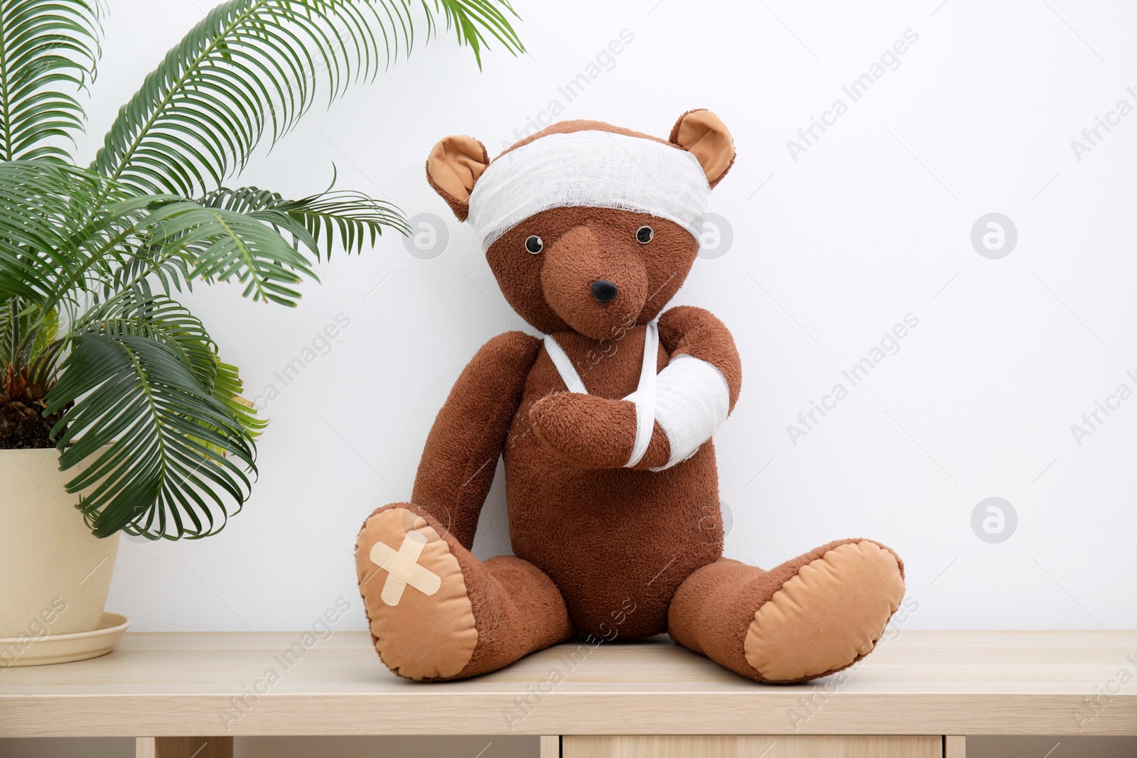 Photo of Toy bear with bandages sitting on wooden shelving unit near white wall