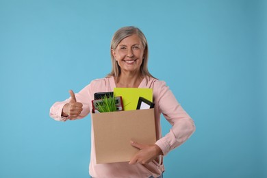 Photo of Happy unemployed senior woman with box of personal office belongings showing thumb up on light blue background