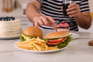 Photo of Overweight man with glass of cold drink taking burger from plate at table in kitchen, closeup