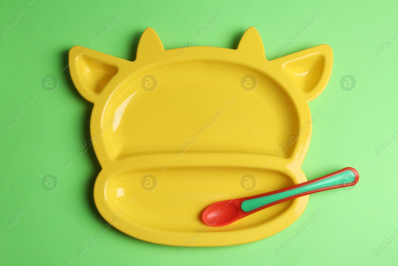 Photo of Cute animal shaped plate and plastic spoon on light green background, top view. Serving baby food