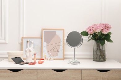 Photo of Mirror, cosmetic products, perfumes and vase with pink roses on white dressing table in makeup room