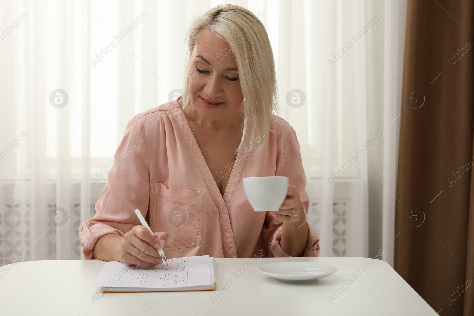 Photo of Middle aged woman with cup of drink solving sudoku puzzle at table indoors