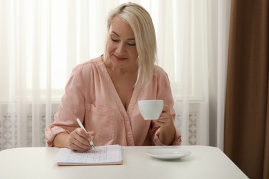 Photo of Middle aged woman with cup of drink solving sudoku puzzle at table indoors