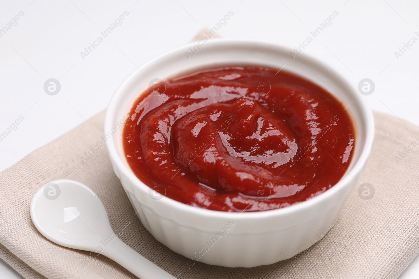 Photo of Organic ketchup in bowl and spoon on white table, closeup. Tomato sauce